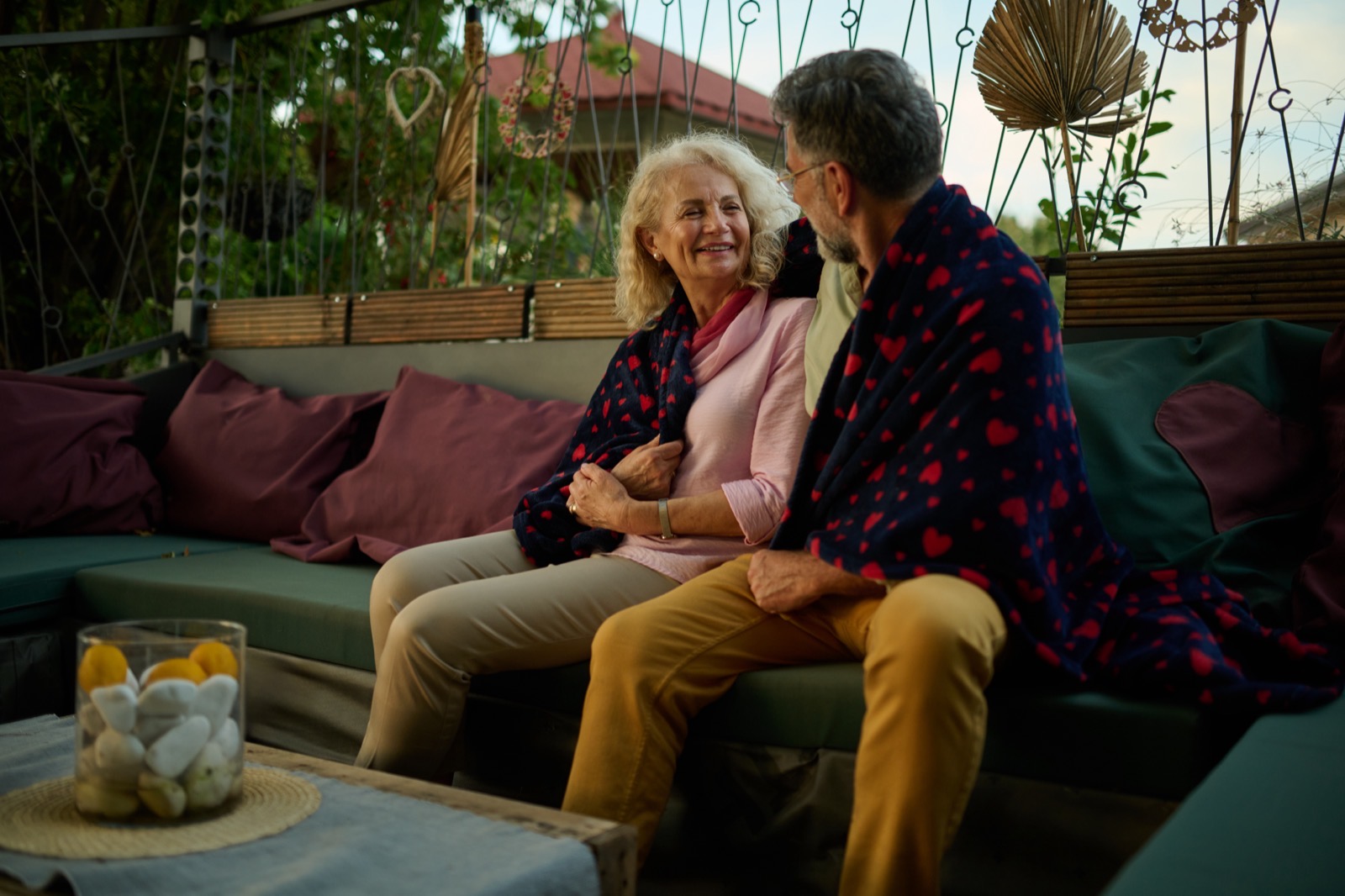Un couple de seniors installé sous un plaid sur la terrasse d’une maison, en fin de saison